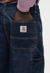 Χέρι με σκούρο δέρμα που έχει μπει στην πίσω τσέπη σκούρου μπλε τζιν παντελονιού Carhartt, με το πορτοκαλί λογότυπο να φαίνεται στην τσέπη.