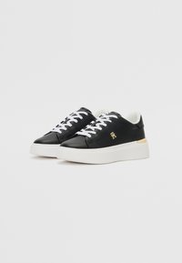 Black leather sneakers with white laces, rounded toe, and a thick white sole. Gold accent on the heel. Smooth texture with minimal design.