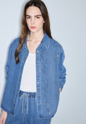 Frau mit langen braunen Haaren, die eine blaue Jeansjacke und passende Hose mit schwarzen gepunkteten Mustern über einem weißen Hemd trägt, steht vor einer schlichten Wand.