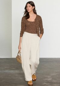 Haut à manches longues marron �à pois, superposé sur un pantalon large taille haute blanc. Le mannequin tient un petit sac à main beige et porte des sandales à talons tan.