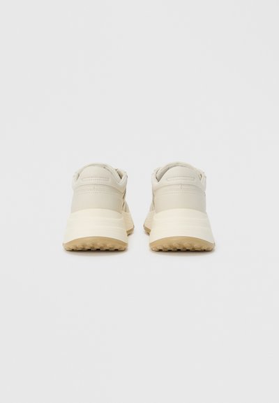 Μπεζ αθλητικά παπούτσια με λείο επάνω μέρος, επενδεδυμένο γιακά και υφασμένο καουτσούκ σόλα με μικρά καρφιά για πρόσφυση. Απλός, μινιμαλιστικός σχεδιασμός.