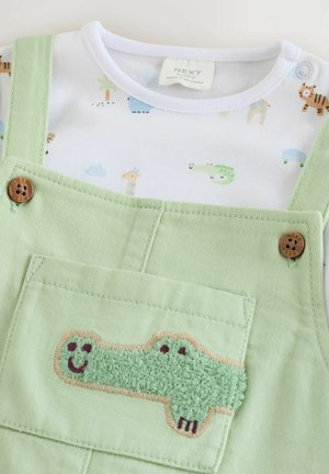 Une salopette vert clair avec des boutons en bois, présentant un design texturé de crocodile sur la poche frontale et une chemise blanche à motifs en dessous.