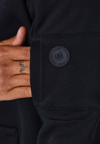Felpa nera con una texture liscia, dotata di un'applicazione rotonda con logo sulla manica. Mano posizionata sul tessuto vicino al logo.