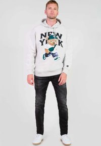 Grijze hoodie met een berenafbeelding en "NEW YORK" tekst, gecombineerd met donkere jeans en witte sneakers met tan kleurige zolen. Soepele textuur.