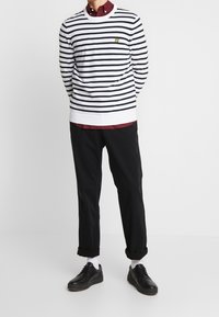 Camicia a maniche lunghe bianca e blu a righe con scollatura rotonda e un piccolo logo giallo, abbinata a pantaloni neri e scarpe nere.