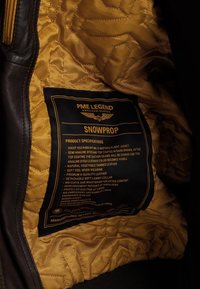 Brun flyjakke med en kviltet gul foring, der har en sort etiket der detaljerer specifikationer og funktioner. Blød læderkonstruktion.