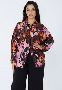 Woman with long dark hair wearing a loose button-up shirt with pink, orange, and brown abstract patterns, paired with black wide-leg pants.
