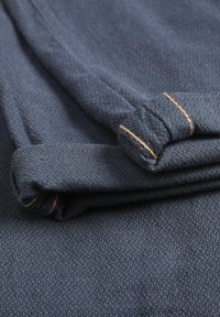 Tmavomodrá látka so subtilným textúrovaným vzorom, vybavená zrolovanými manžetami a kontrastným oranžovým prešitím.