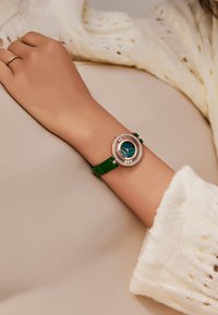 Reloj de pulsera redondo con correa de cuero verde y un dial azul, enmarcado por un bisel de oro rosa texturizado adornado con pequeñas gemas.