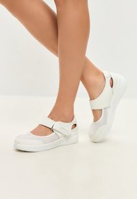 White athletic shoes with mesh panels, a wide strap with a Velcro closure, rounded toe, and a cushioned wedge sole.