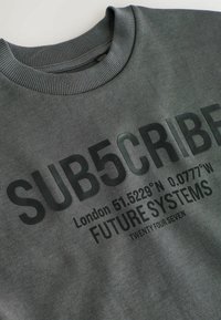 Sivi dukserica s rebrastim ovratnikom. Na prednjoj strani se nalaze crni natpisi: "SUB5CRIBE," koordinate, "FUTURE SYSTEMS," i "TWENTY FOUR SEVEN."