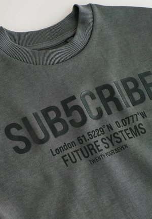 Sivá mikina s rebrovým golierom, so silným čiernym textom: "SUB5CRIBE", súradnicami a "FUTURE SYSTEMS" v matnom prevedení.