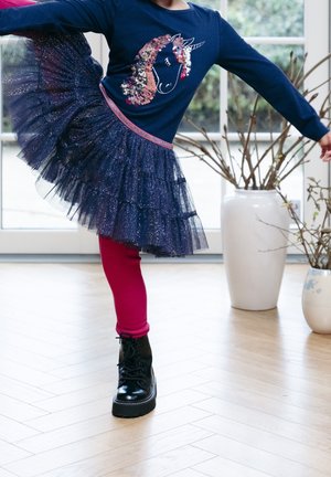 Enfant portant un pull bleu marine avec un motif licorne, un tutu bleu marine pailleté, des leggings roses et des bottes noires, posant à l'intérieur près de grandes fenêtres et de vases.