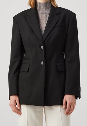 Person iført en sort skræddersyet blazer over en højhalset ternet top, kombineret med løse hvide bukser, vist fra nakke til midt på låret.