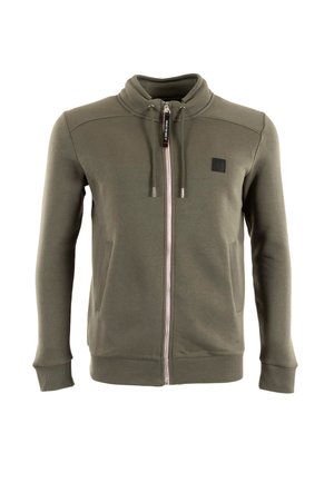 Chaqueta de color verde oliva con cremallera y cuello alto, bolsillos frontales, cordones, puños y dobladillo acanalados, y un pequeño parche negro en el pecho.