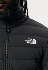 Casaco acolchoado preto com gola alta, apresentando um logo branco no peito e fecho com zip. Tecido liso com um aspeto texturado.