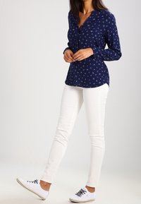 Camicia a maniche lunghe di colore blu navy con motivo, chiusura a bottoni, abbinata a pantaloni skinny bianchi e sneakers bianche. La camicia ha un collo a V.