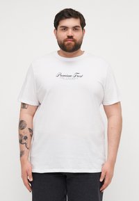 White cotton t-shirt features short sleeves, a crew neck, and a centered black text logo. Paired with dark gray pants.