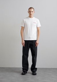 Camiseta de algodón blanca con "THE COMING" impreso en rojo, combinada con jeans de mezclilla negros de corte holgado y zapatos negros. Diseño simple y ajuste casual.