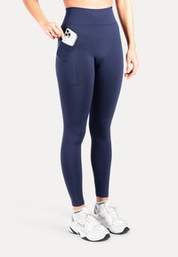 Marineblauwe leggings met hoge taille, een zijzak en een gladde, rekbare stof. Gecombineerd met witte sneakers met zwarte accenten.