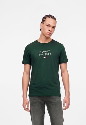 Jeune homme aux cheveux tressés portant un t-shirt Tommy Hilfiger vert foncé et un jean gris, debout devant un fond blanc uni.