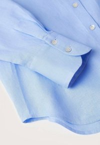Jasnoniebieska koszula o gładkiej fakturze, z mankietem na rękawie i dwoma srebrnymi guzikami. Dolna krawędź ma lekko zaokrąglony kształt.
