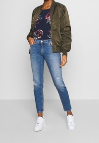 Giacca bomber verde oliva sopra una maglietta floreale blu navy, abbinata a jeans chiari strappati e sneakers bianche. Texture e colori semplici.