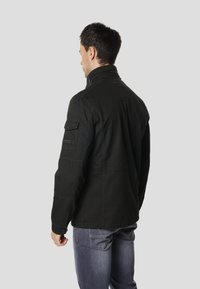 Černá bavlněná bunda s vysokým límcem, manžetami na knoflíky a boční kapsou na levém rukávu. Nošena přes modré džíny.