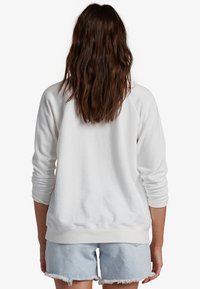 Kvinna med långt, brunt, vågigt hår som bär en vit sweatshirt och ljusblå fransade denimshorts, stående med ryggen mot kameran.