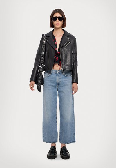 Femme portant une veste en cuir noire, un crop top noir avec imprimé cerises, un jean large bleu clair, des mocassins noirs, des lunettes de soleil et un sac à épaule noir.