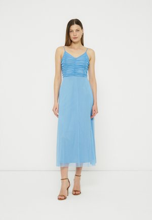 Γυναίκα που φοράει ένα αμάνικο γαλάζιο midi φόρεμα με ραμμένο μπούστο και λεπτές τιράντες, συνδυασμένο με καφέ πέδιλα με λουράκι στον αστράγαλο και ψηλό τακούνι.