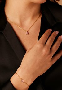 Gouden ketting en armband set met roze bloemenpendanten, gladde textuur, gedragen tegen een zwarte top.