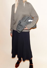 Pull à col roulé en maille grosse torsade gris, jupe mi-longue noire, tenant une veste matelassée grise, et chaussures marron avec des chaussettes noires.