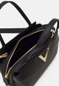 Svart texturerad läderhandväska med gulddragkedja, dubbla handtag och en stor dekorativ guldfärgad detalj. Inuti finns flera fack.