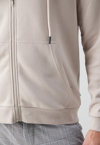 Felpa leggera beige con zip, tasca a marsupio, orlo a costine e cordini. Indossata sopra pantaloni grigi a quadri. Texture liscia.