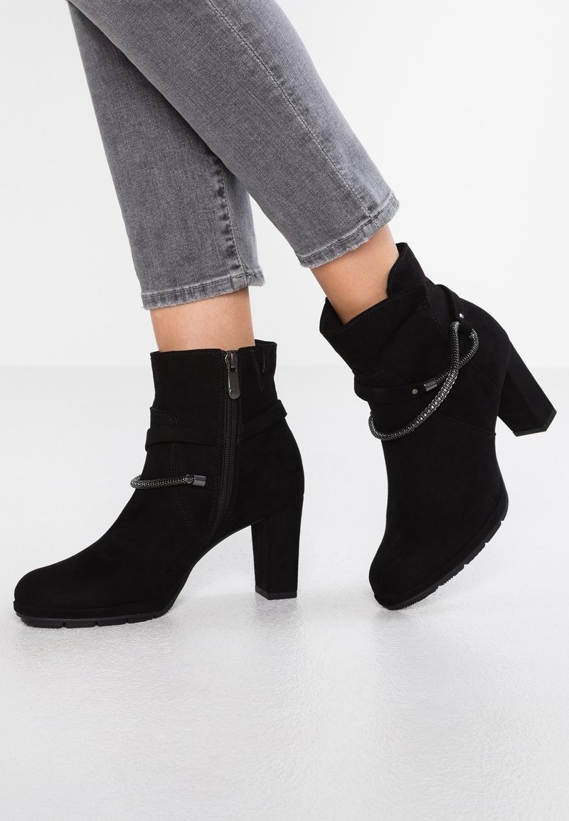 Bottines noires en daim avec talons blocs et fermetures éclair latérales portées avec un jean gris raccourci sur un sol blanc.