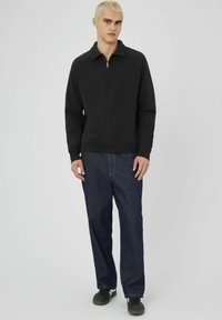 Jeune homme aux cheveux blonds portant un sweat-shirt noir à fermeture éclair, un jean bleu foncé et des baskets noires, se tenant contre un fond uni.