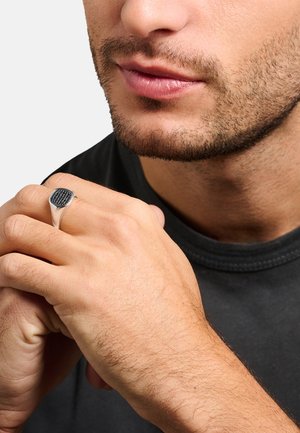 Primo piano di un uomo con una leggera peluria sul viso che indossa un anello d’argento con pietre nere sulla mano destra, mani unite vicino al volto.