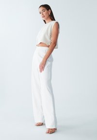 Hvid cropped top med sidebindinger, parret med brede, hvide teksturerede bukser. Modellen har beige sandaler på og en minimalistisch ørering.