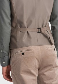 Zweifarbige Bekleidung mit einem taupefarbenen Rücken, ausgestattet mit einem Gürtel in der Taille und strukturierten grauen Ärmeln. Cord-Hosen in einem hellen Beige mit Gesäßtaschen.