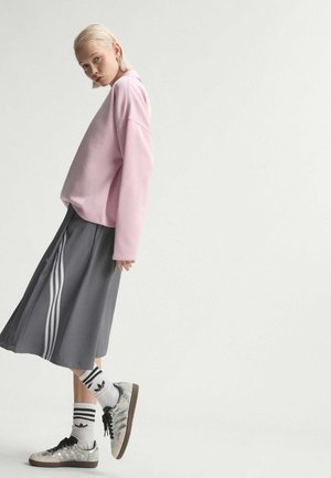 Jeune personne aux cheveux blonds courts portant un sweat-shirt rose, une jupe grise à rayures blanches, des chaussettes blanches rayées et des baskets argentées, debout sur une jambe.