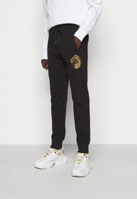 Schwarze Jogginghose mit einem goldenen Emblem am linken Bein, ausgestattet mit einem Kordelzug an der Taille und seitlichen Taschen, kombiniert mit weißen, klobigen Sneakern.