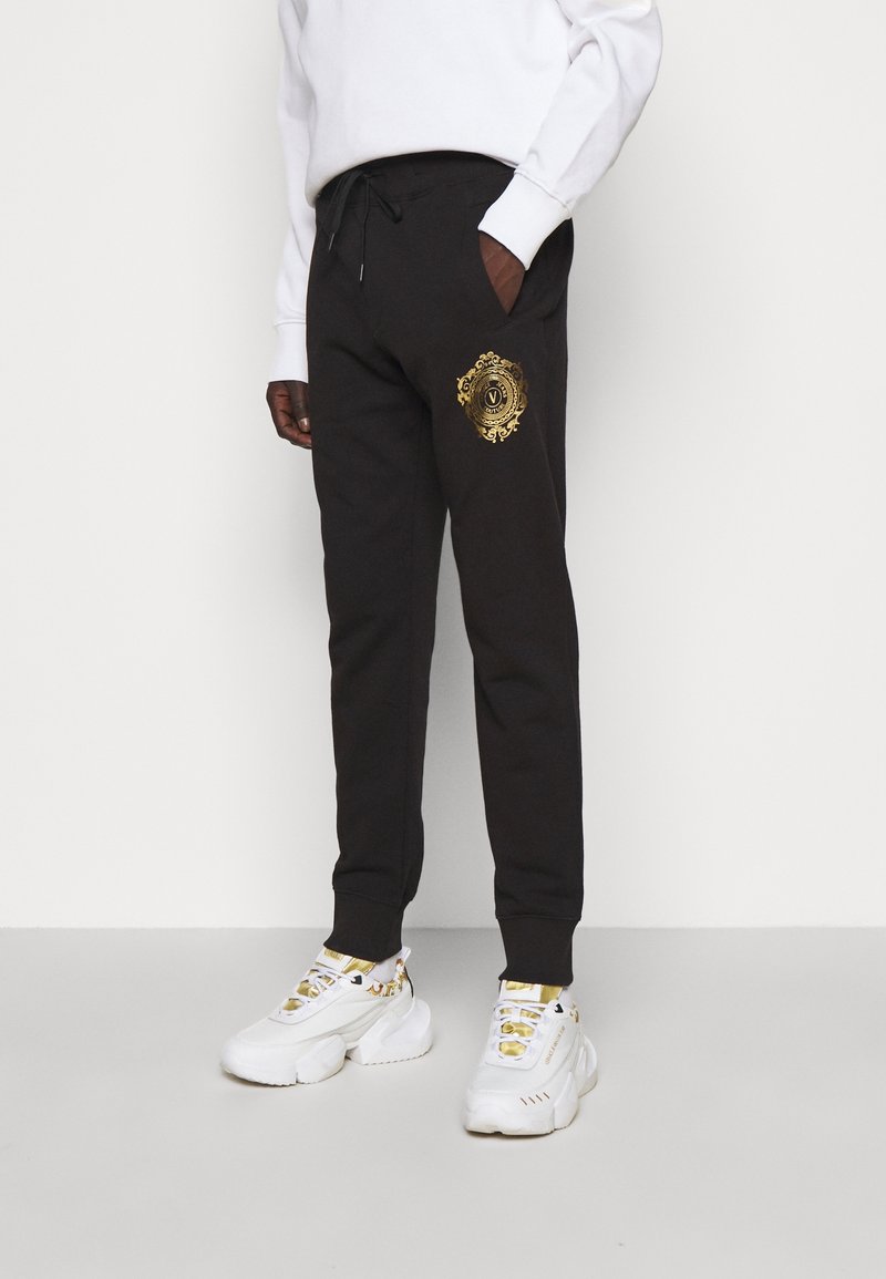 Schwarze Jogginghose mit einem goldenen Emblem am linken Bein, ausgestattet mit einem Kordelzug an der Taille und seitlichen Taschen, kombiniert mit weißen, klobigen Sneakern.
