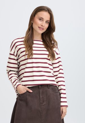 Jeune femme aux longs cheveux bruns portant un haut à manches longues blanc et bordeaux à rayures et une jupe marron foncé, la main dans la poche.