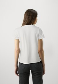 Biała bawełniana koszulka z krótkim rękawem, okrągłym dekoltem i luźnym krojem. Tył jest gładki, zestawiona z ciemnoniebieskimi dżinsami.