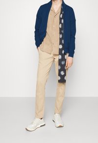 Marinblå zip-up cardigan, beige mockaskjorta, beige byxor, mönstrad blå scarf och vita sneakers med beige detaljer. Avslappnad outfit.