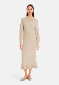 Strickkleid in hellem Beige mit einem Rüschen-Ausschnitt, langen Ärmeln, gerippter Textur und einem ausgestellten, gewellten Saum. Kombiniert mit beigen Ballerinas.
