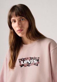 Sweatshirt rose clair avec un grand logo Levi's noir et floral à l'avant. Le matériau semble doux avec une coupe décontractée.