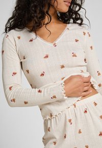 Lange mouw top in gebroken wit met een gestructureerde stof en teddybeer patroon, bevat een gegolfde V-halslijn en gerimpelde manchetten.