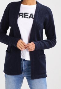 Navyblauer Cardigan aus weichem, strukturiertem Stoff, mit offenem Vorderteil, langen Ärmeln und zwei vorderen Taschen, getragen über einem weißen Hemd.